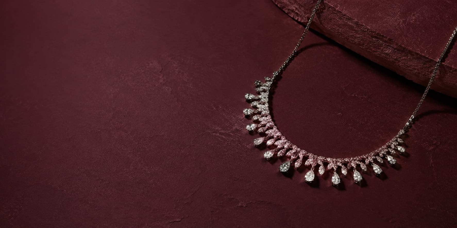 A silver necklace with pear-drop stones resting on a deep burgundy stone surface.
